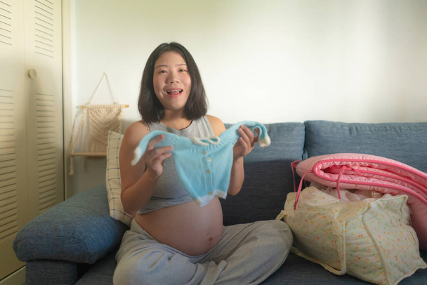 young happy and beautiful Asian Chinese woman pregnant on couch enjoying playful with little baby clothes smiling relaxed at home in pregnancy and maternity concept - Foto, immagini