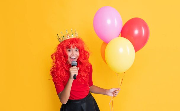 glad child in crown with microphone and party balloon on yellow background. - Foto, afbeelding