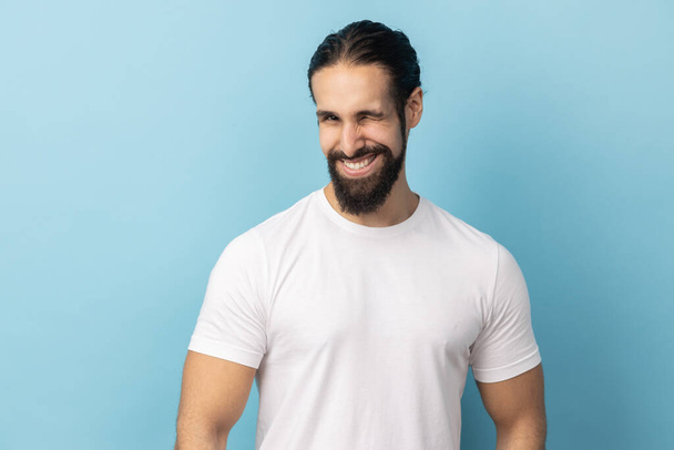 Portrait of cheerful man with beard wearing white T-shirt being in good mood, smiling broadly and winking at camera with toothy smile. Indoor studio shot isolated on blue background. - Fotografie, Obrázek