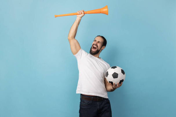 Portrait of man with beard wearing white T-shirt cheering for favorite football team, holding horn and ball in hands, screaming happily. Indoor studio shot isolated on blue background. - Photo, Image