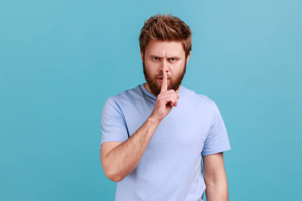 Portret van een man met een baard die zijn vinger in de buurt van lippen houdt, die zijn gebaar toont en vraagt om te zwijgen en heel, privacy te zijn, kijkend naar de camera. Indoor studio schot geïsoleerd op blauwe achtergrond. - Foto, afbeelding