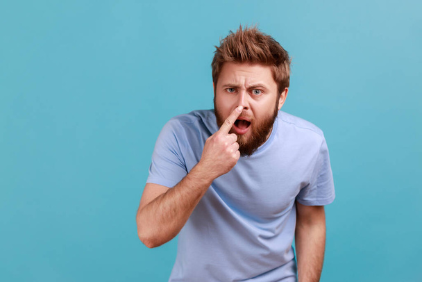 You are liar. Portrait of shocked bearded man standing, touching his nose and showing lie gesture, suspecting falsehood, dishonest talk. Indoor studio shot isolated on blue background. - Photo, Image