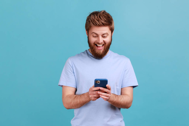 Portrait of happy excited bearded man using online service on mobile phone and expressing pleasant emotions, texting messages for friends. Indoor studio shot isolated on blue background. - Foto, Bild