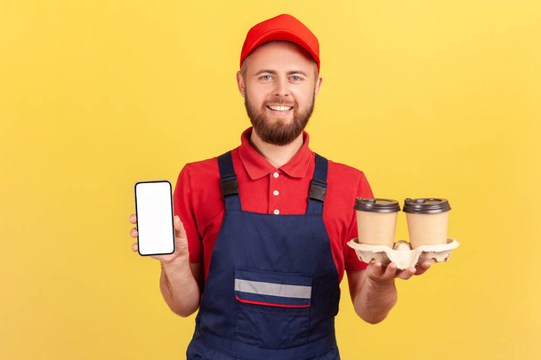Portrait of courier man in blue overalls and red cap, holding takeaway coffee and showing cell phone with blank screen for advertisement. Indoor studio shot isolated on yellow background. - Foto, immagini
