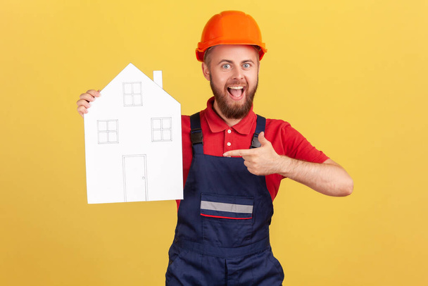 Portrait of builder man pointing at paper house, help in relocation, home repair services, looking at camera with excitement, wearing overalls and cap. Indoor studio shot isolated on yellow background - 写真・画像
