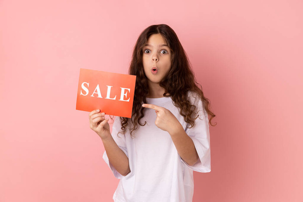 Portrait of surprised shocked little girl wearing white T-shirt pointing at card with sale inscription, looking at camera with open mouth. Indoor studio shot isolated on pink background. - Foto, afbeelding