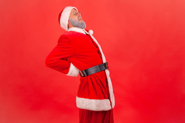 Sick elderly man with gray beard wearing santa claus costume feeling sharp pain in kidneys, back injury, pinched sciatic nerve, suffering. Indoor studio shot isolated on red background. - Foto, afbeelding