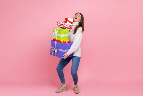 Full length portrait of happy brunette woman holding heavy stack of presents, celebrating holiday, wearing white casual style sweater. Indoor studio shot isolated on pink background. - Photo, image
