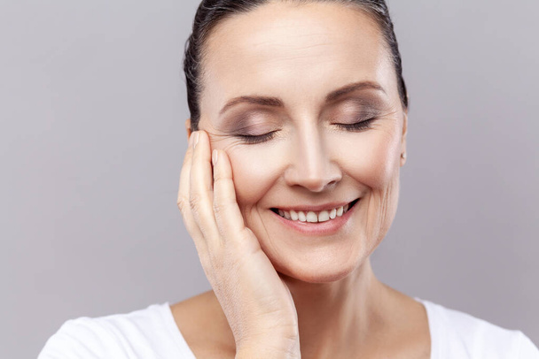 Closeup portrait of middle aged woman gently touching face with one hand and smiling, having healthy pure skin, female enjoying spa facial treatment. Indoor studio shot isolated on gray background. - Foto, afbeelding