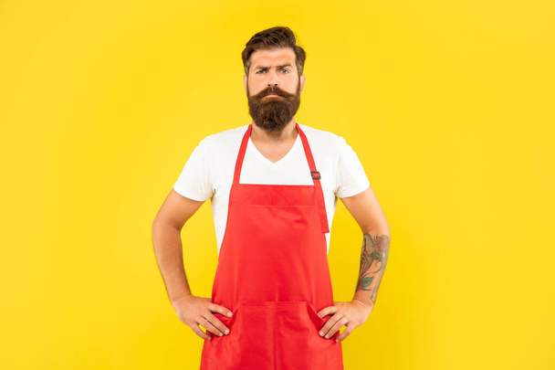 Confident serious man with arms akimbo in red apron yellow background, household. - Фото, изображение
