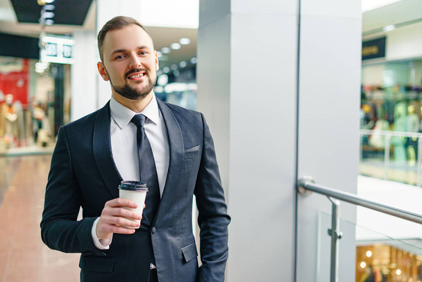 A young bearded man dressed in a business suit at the mall with a cup of coffee. A young man in an elegant business suit strolls through a mall drinking coffee as he goes - Φωτογραφία, εικόνα
