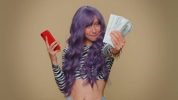 Happy young woman with lush wig looking smartphone display sincerely rejoicing win, receiving money dollar cash banknotes, success lottery luck. Adult stylish female girl on beige studio background - Foto, Bild