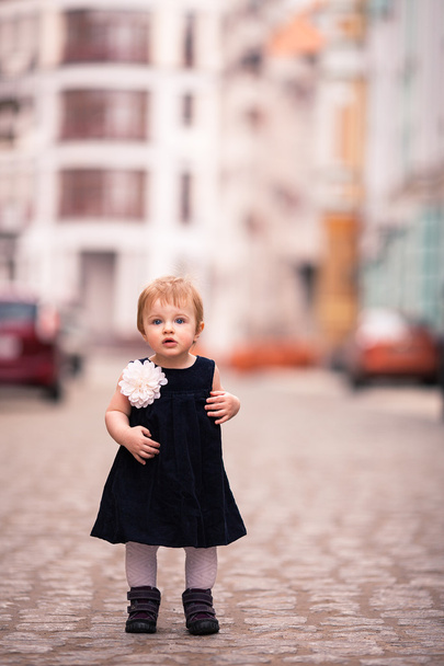 Een babymeisje in donkere jurk met grote witte bloem staat op de straat - Foto, afbeelding