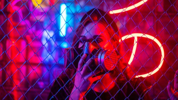 Caucasian woman in a gas mask behind a fence in a neon studio - Fotoğraf, Görsel