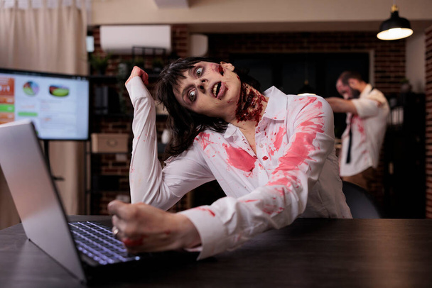 Horror cruel zombie using laptop at desk, undead corpse trying to work on computer in startup office. Creepy aggressive brain eating monster looking terrifying and horrible, sinister danger. - 写真・画像