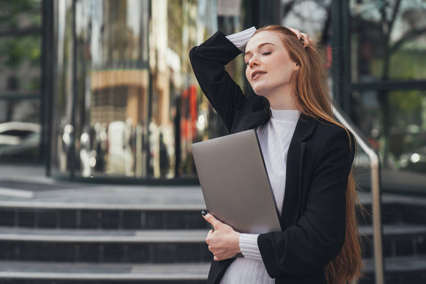 Beautiful redhead woman entrepreneur standing outdoors with laptop in hands, waiting for someone looking aside. Red haired secretary woman. Office work outfit - Fotoğraf, Görsel