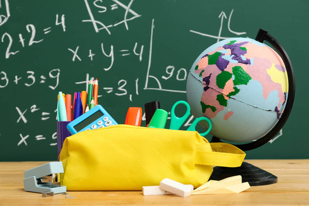 Yellow pencil case with school stationery and globe on table near green chalkboard - Photo, Image