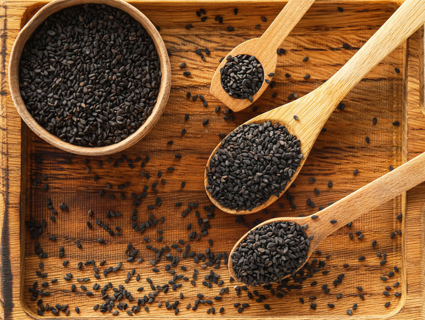 Wooden board with bowl and spoons of black sesame seeds, closeup - Foto, afbeelding