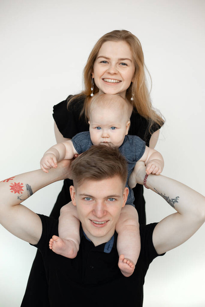 Portrait of young smiling family in dark clothes with plump grey-eyed baby infant toddler standing on white background. Child sitting on fathers shoulders. Family, relationship, having fun concept. - Foto, afbeelding