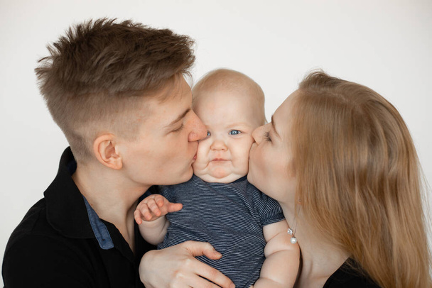 Portrait of young happy attractive family in dark clothes standing with little plump grey-eyed baby infant on white background. Parents kissing child together. Family, relationship, having fun. - Zdjęcie, obraz