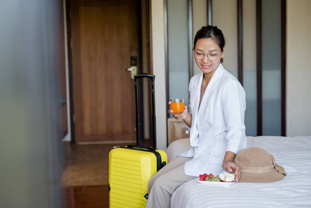 Asijská podnikatelka sedí na posteli v hotelovém pokoji, vyčerpaná po služební cestě, připravená dostat od hotelu ovocnou svačinku, zařízená pro VIP hosty. - Fotografie, Obrázek