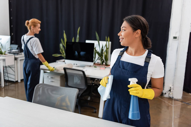 happy bi-racial woman with detergent and rag looking at colleague cleaning office on blurred background - Photo, Image