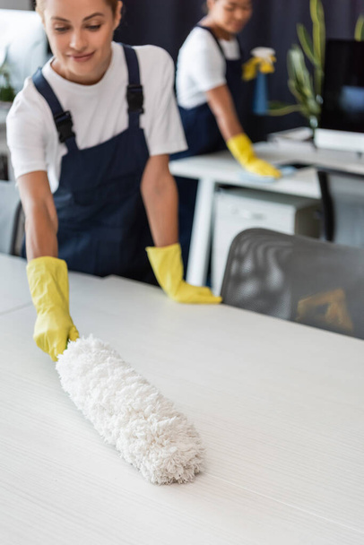 mulher sorridente secretária de limpeza com escova de poeira perto borrada colega bi-racial - Foto, Imagem