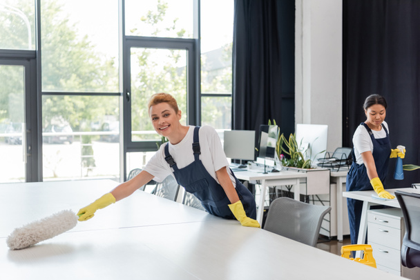 mujer feliz en overoles escritorio de la oficina de limpieza con cepillo de polvo cerca de colega bi-racial en el fondo - Foto, Imagen