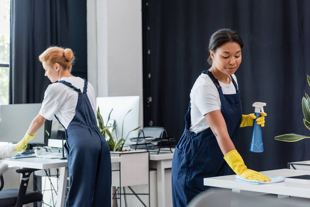 interracial women in overalls cleaning office with dust brush and rag - Photo, Image
