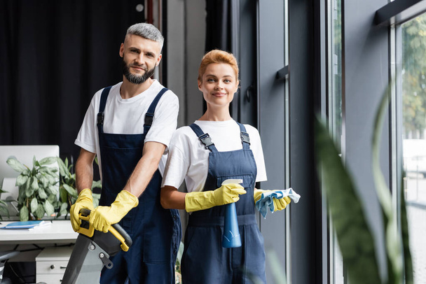 smiling professional cleaners in uniform looking at camera in office - Photo, Image