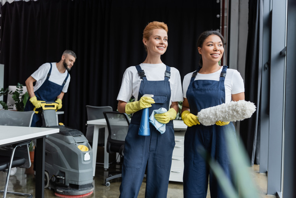 mulheres inter-raciais felizes com suprimentos de limpeza olhando para longe perto do homem com máquina de limpeza de piso - Foto, Imagem