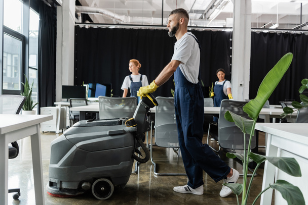 hombre barbudo con máquina de fregar piso eléctrico cerca de limpiadores multiétnicos que trabajan en la oficina - Foto, Imagen