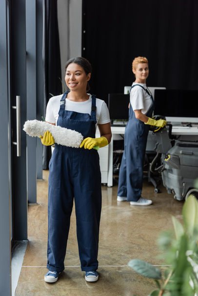 alegre bi-racial mujer en ropa de trabajo de pie con cepillo de polvo cerca de colega con máquina de fregar piso - Foto, Imagen