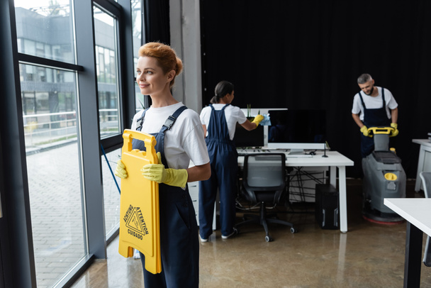 smiling woman holding attention board near windows while interracial colleagues cleaning office - Photo, Image