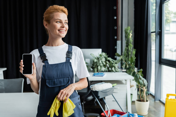 cheerful woman in overalls holding smartphone with blank screen while looking away in office - Photo, Image