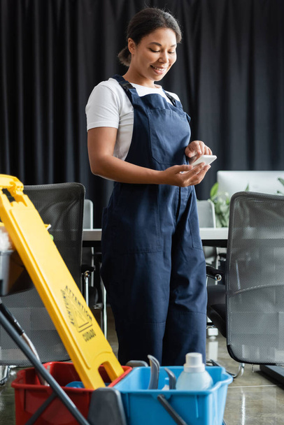 cheerful bi-racial woman in workwear using smartphone near cart with cleaning supplies - Photo, Image