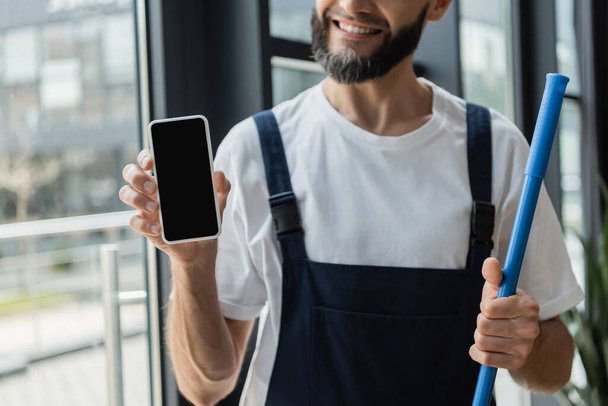 vue partielle du nettoyeur barbu heureux en salopette tenant le téléphone portable avec écran blanc - Photo, image