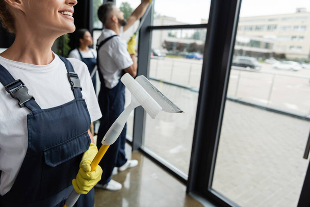 smiling woman in overalls holding window squeegee near blurred multiethnic colleagues - Photo, Image