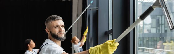 homme barbu laver les fenêtres de bureau près des femmes multiculturelles floues, bannière - Photo, image