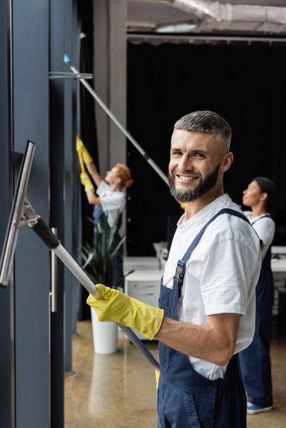 bearded man looking at camera while washing windows near multiethnic colleagues on blurred background - Photo, Image