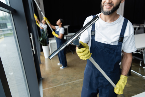 smiling man holding window squeegee near blurred interracial women - Photo, Image