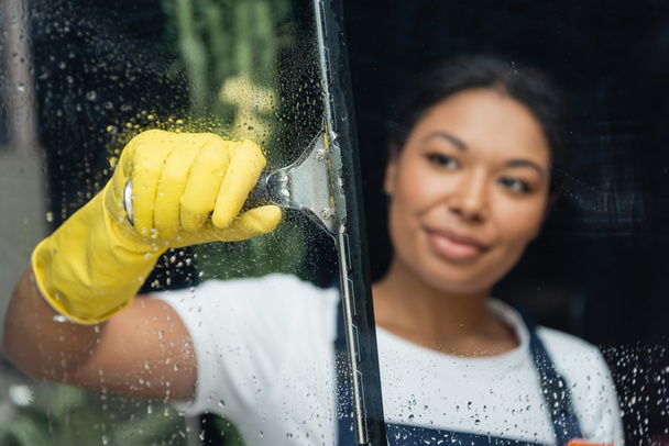 blurred bi-racial woman in rubber glove cleaning glass with window wiper - Photo, Image