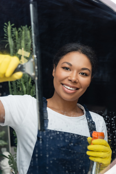 bi-racial happy woman in rubber gloves looking at camera while washing window in office - Photo, Image