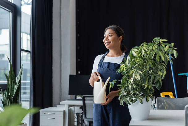 happy bi-racial woman in workwear standing with watering can near plant and looking away - Photo, Image