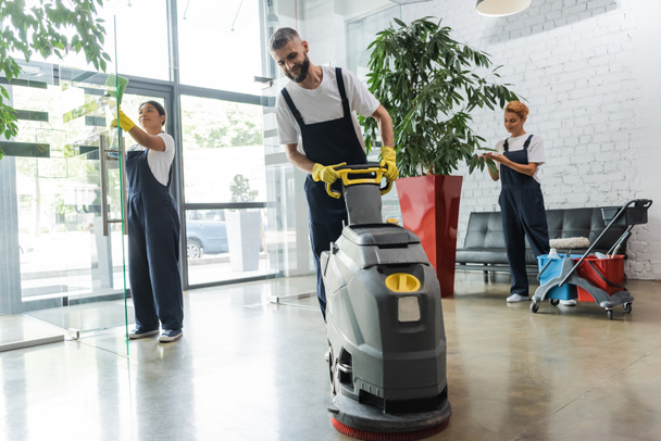 man in workwear with floor scrubber machine near interracial women cleaning office lobby - Photo, Image