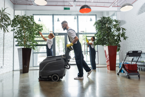 side view of man in overalls with electrical floor scrubber machine near interracial colleagues - Foto, Bild