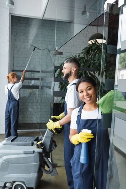 mujer bi-racial sonriente con detergente mirando a la cámara cerca de colegas de limpieza de la oficina - Foto, imagen