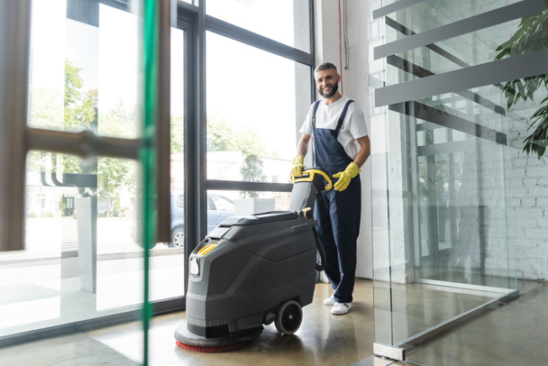 Professioneller Reiniger in voller Länge mit elektrischem Bodenwäscher, der in die Kamera lächelt - Foto, Bild