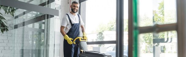 limpiador profesional sonriendo a la cámara cerca de la máquina de fregar piso, pancarta - Foto, Imagen