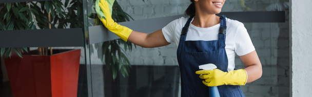 vue recadrée de la femme bi-raciale souriante dans des gants en caoutchouc nettoyage porte en verre dans le bureau, bannière - Photo, image
